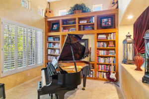 Music and reading room at the West Valley Estate featuring a grand piano, custom wood bookshelves, natural light from plantation shutters, and eclectic decor.